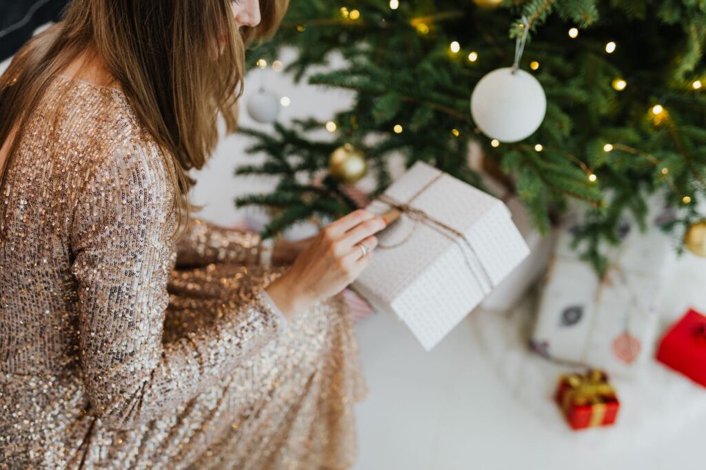 Mujer con un vestido elegante y brillante abriendo un regalo de Navidad frente al árbol de navidad. El desafío de la Concepción en Navidad.
