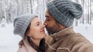 A couple with woolly hats on kissing in a snowy forest