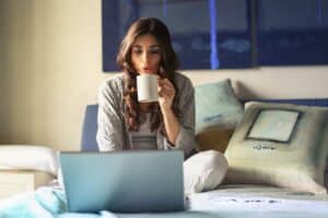 Woman drinking hot coffee on bed while looking at laptop